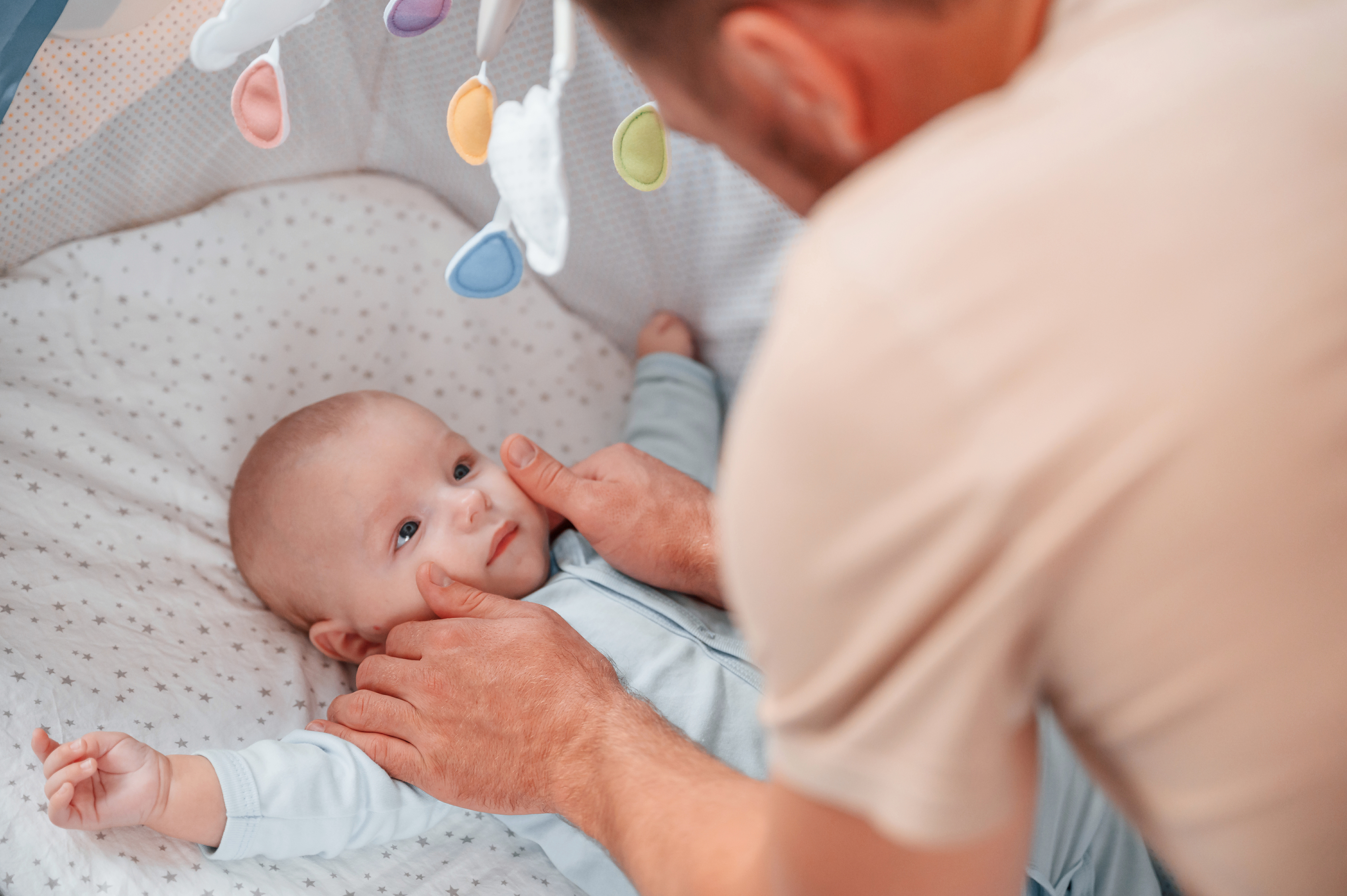 Parent soothing an alert baby beside a clear crib
