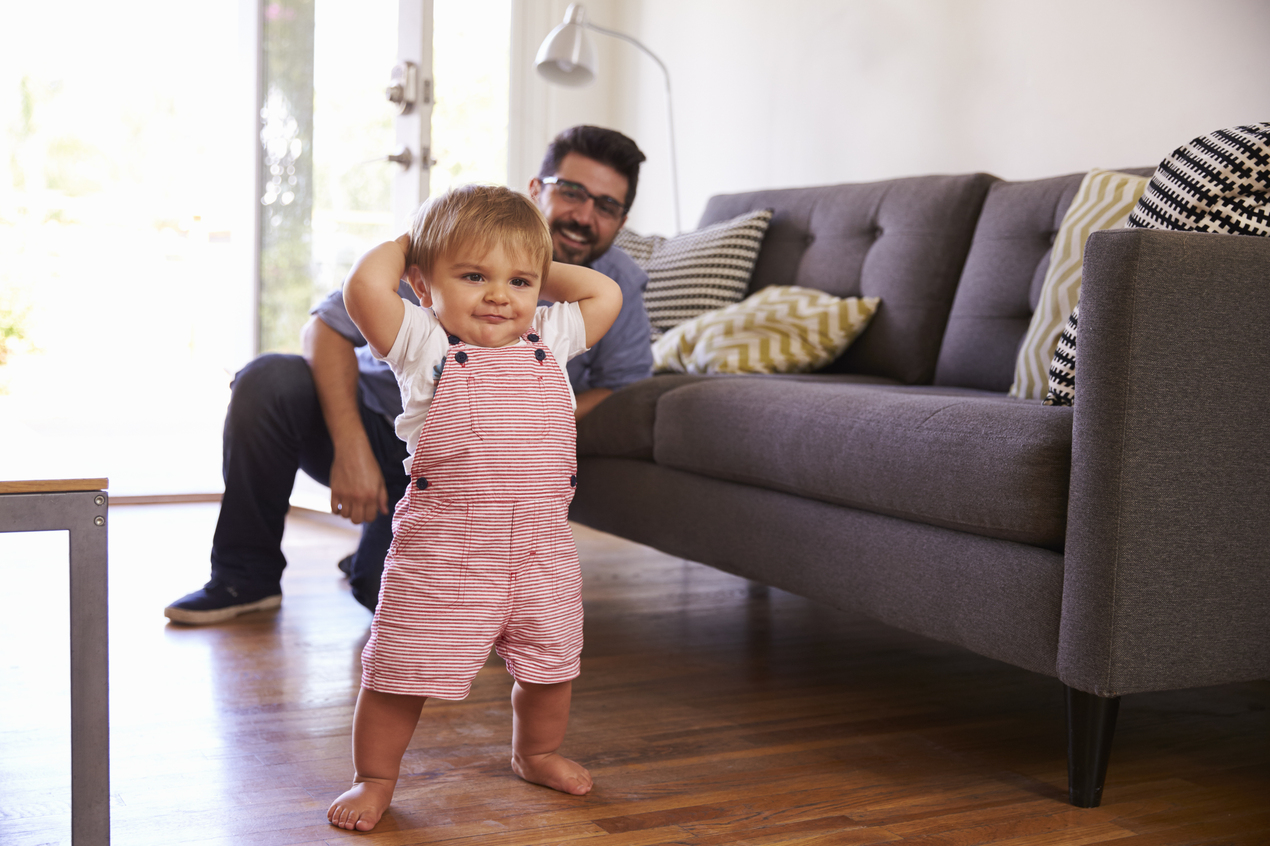 Toddler taking first steps in a living room while a parent watches